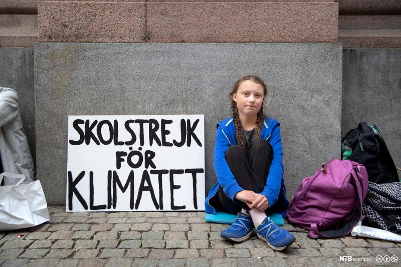 Greta Thunberg sittende utenfor det svenske parlamentet. Til venstre for henne står en plakat med teksten "Skolstrejk för klimatet". Til høyre for henne en lilla og en sort skolesekk og et pledd. Bildet er tatt i august 2018. Foto.