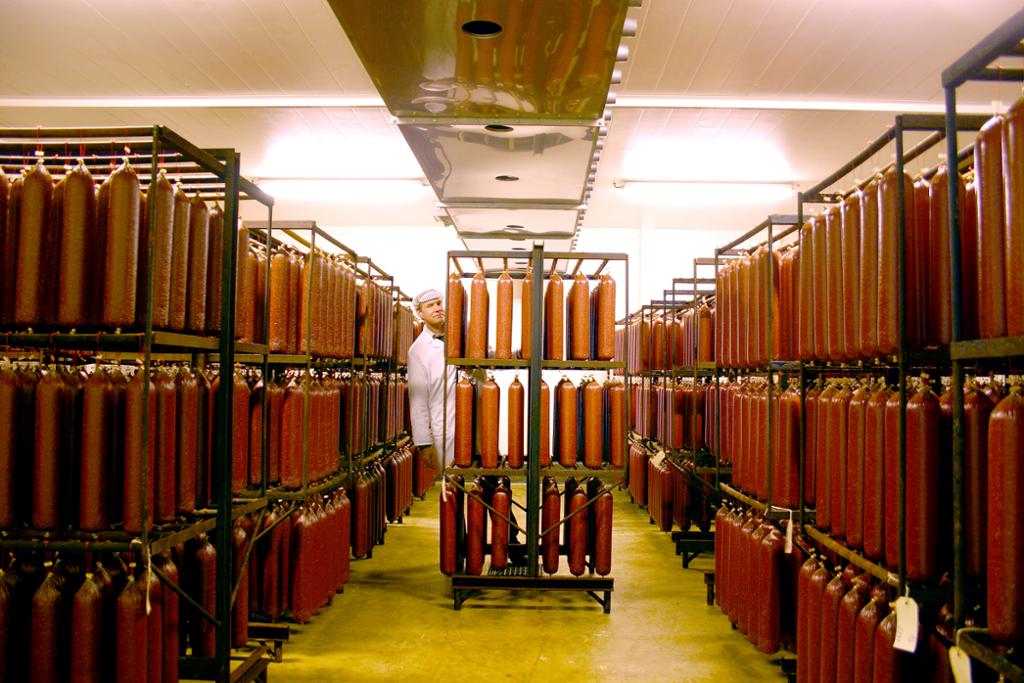 En pølsemaker står inne i en hall med rekker av spekepølser som henger på stativer. Foto.