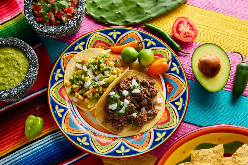 Et bord med en fargerik duk. Oppå bordet står en tallerken med meksikanske tacos, én med kjøttfyll og én med vegetarisk fyll. Guacamole og tomatsalsa i skåler, avokado, chili og tomat ligger ved siden av. Foto.