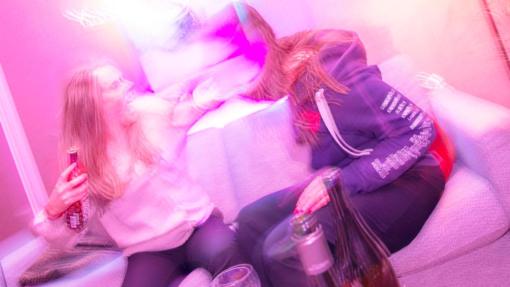 To jenter på fest slåst i ein sofa. Det er flasker med alkohol i nærleiken. Foto.