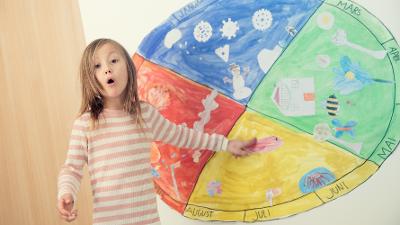 Ei lita jente står med et overrasket blikk og peker på en fargerik plakat med illustrasjoner av årstidene. Foto.