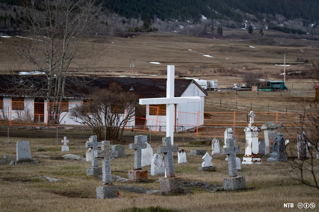 Photo: We see a white building with boarded-up windows. Outside are gravestones and a large, white cross. 