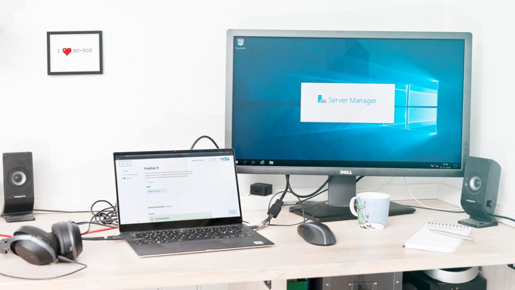 Arbeidspult med headset, mus, bærbar PC, kaffekopp og ekstern skjerm. Skjermene viser ndla.no og Windows Server Manager. Foto.