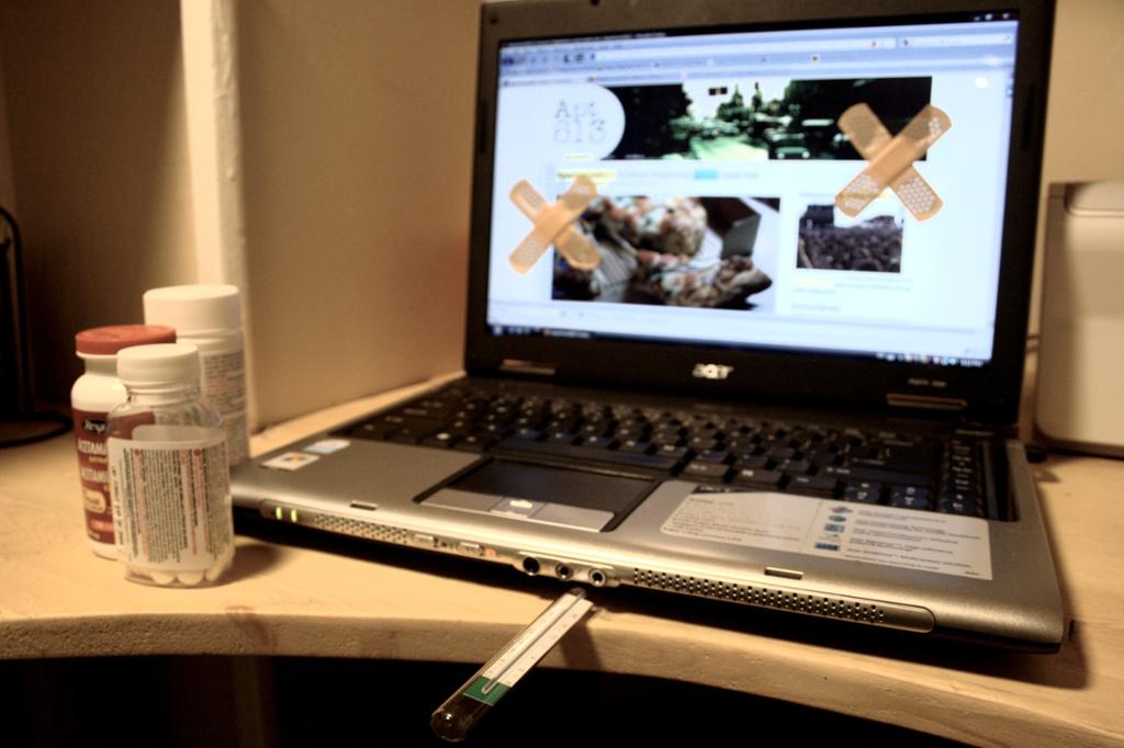Datamaskin med plasterlapper på skjermen. Et termometer ligger under tastaturet. Tre pilleglass i forgrunnen. Foto.