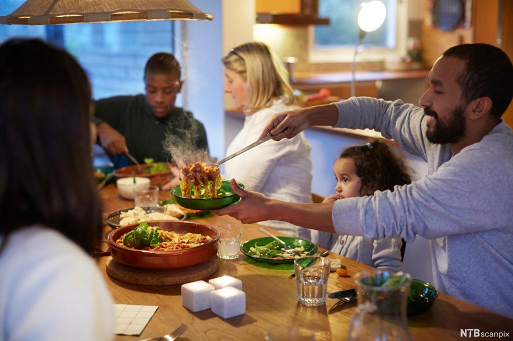 Familie med mor, far og barn i ulik alder spiser middag på et kjøkken. Far øser opp i tallerkenene til barna. Maten består av pasta, kjøtt og grønnsaker. Foto.