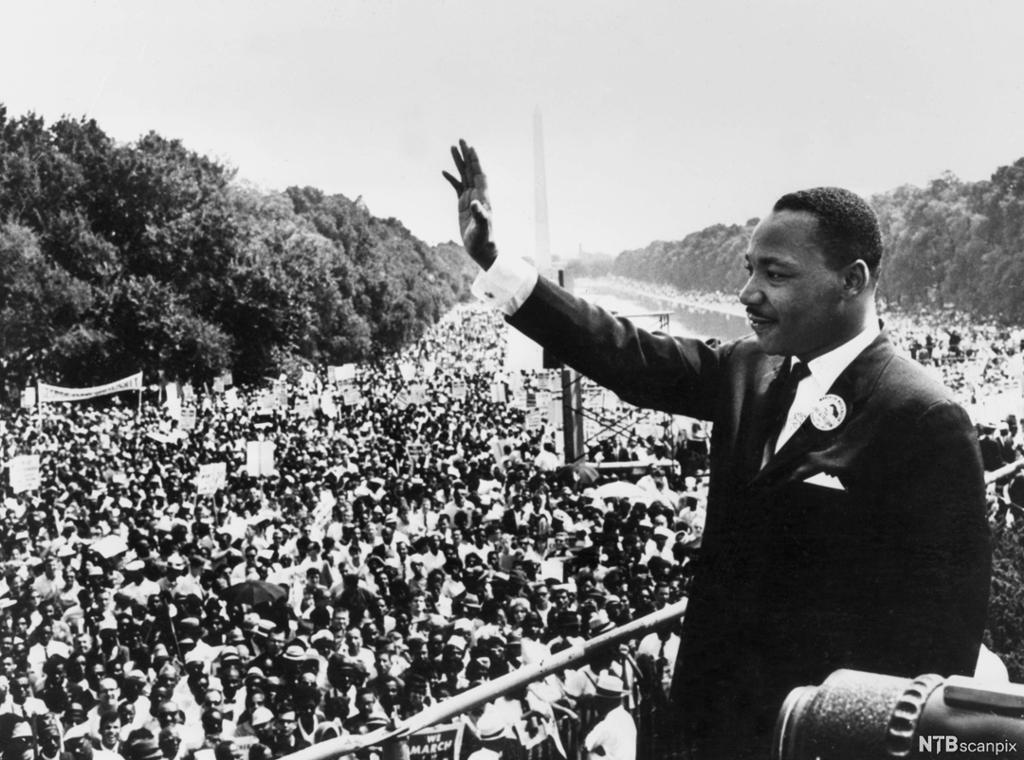 Photo: Martin Luther King Jr. waves at the crowd in Washington. We see thousands of people crammed together. In the background, we see the Washington memorial. 