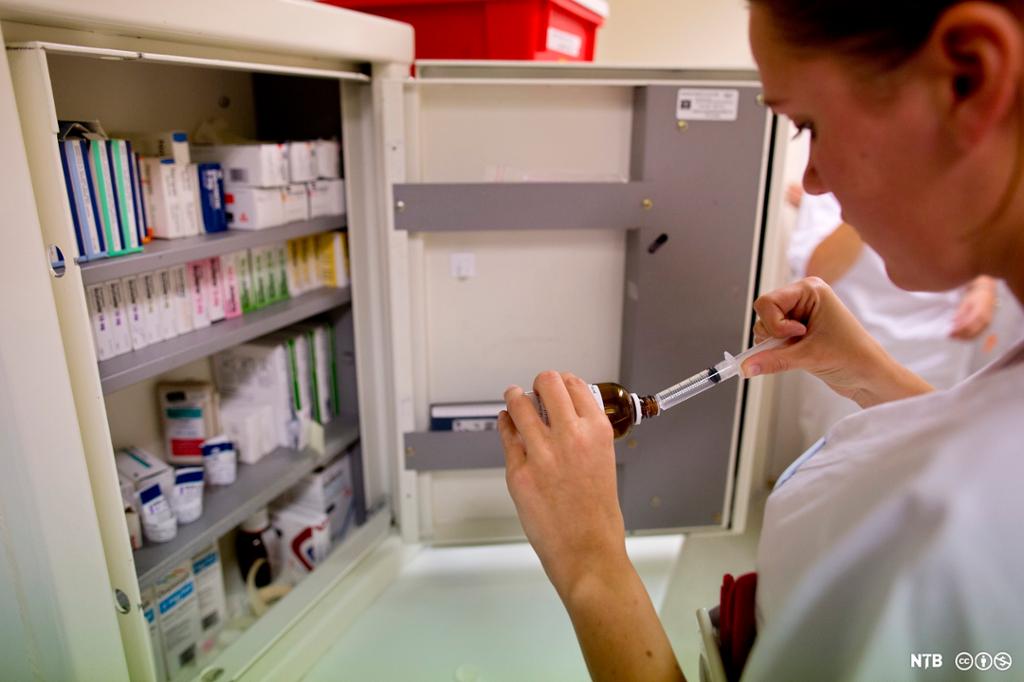 Kvinnelig sykepleier trekker opp medisin fra ei flaske i medisinskap. Foto. 