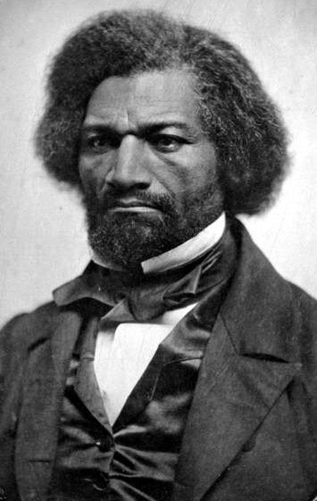 A black and white photo of Fredrick Douglass. He is a Black man dressed in a suit and a bow. He has thick hair and a beard. His expression is sad, or even angry.