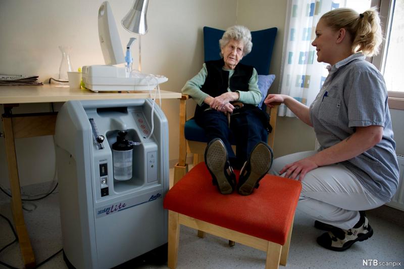 Pasient og pleier på en helseinstitusjon. En eldre kvinne sitter i en stol, en helsefagarbeider sitter på huk ved siden av henne. Til venstre står det medisinske apparater. Foto. 