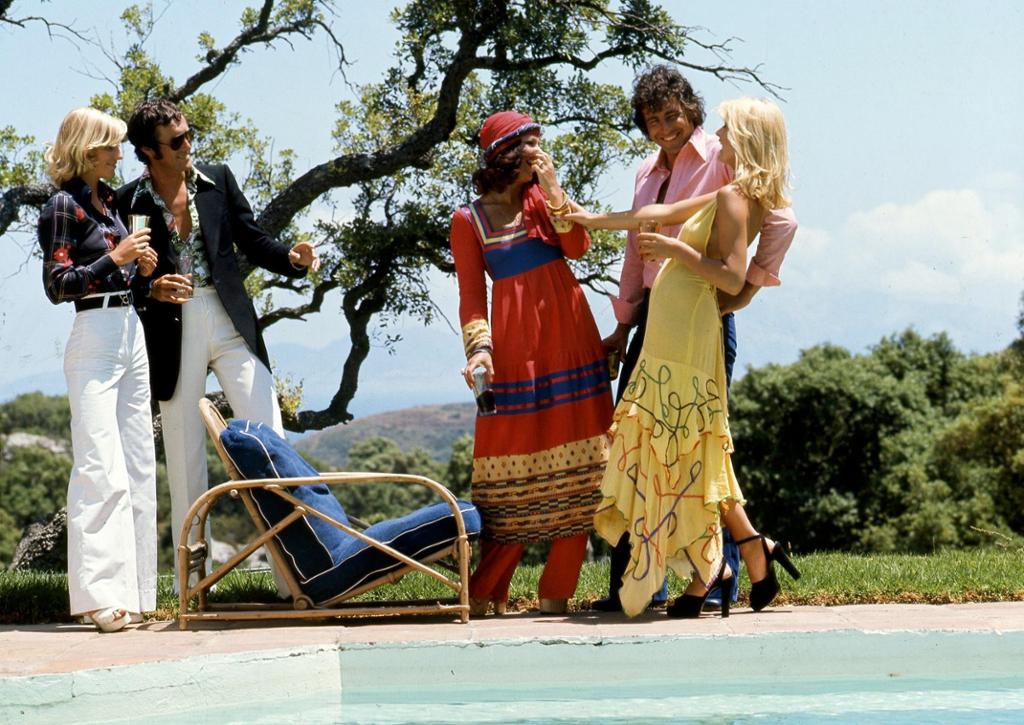 Fem unge mennesker med klær fra 70-tallet ved en solstol ved en bassengkant. En kvinne med hvit slengbukse med høyt liv og mørk bluse med blomstermønster. En mann med hvit bukse, blomstrete skjorte undre svart dressjakke og solbriller. En kvinne med rød og mønstrete kjole med matchende hodeplagg. En mann med blå bukse og rosa skjorte. En kvinne med gul kjole. Foto.