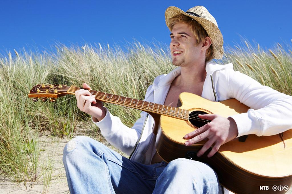 En ung mann med lyse klær sitter ute med gitar og hatt. Foto.