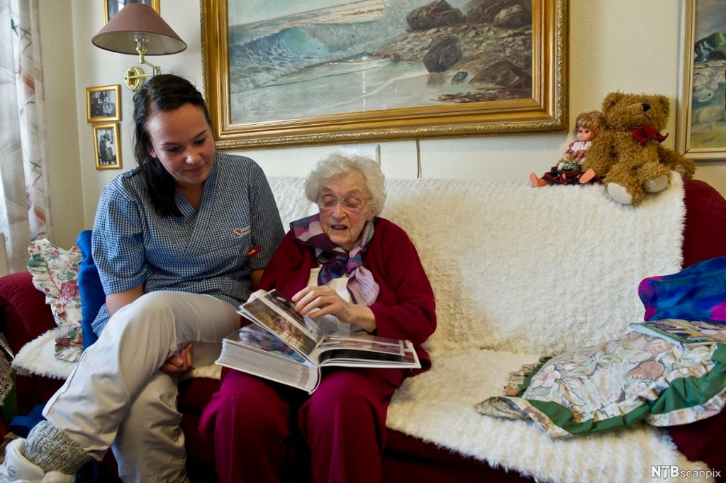 Pleier og eldre beboer på en institusjon sitter ved siden av hverandre i en sofa og ser på et fotoalbum sammen. Foto.