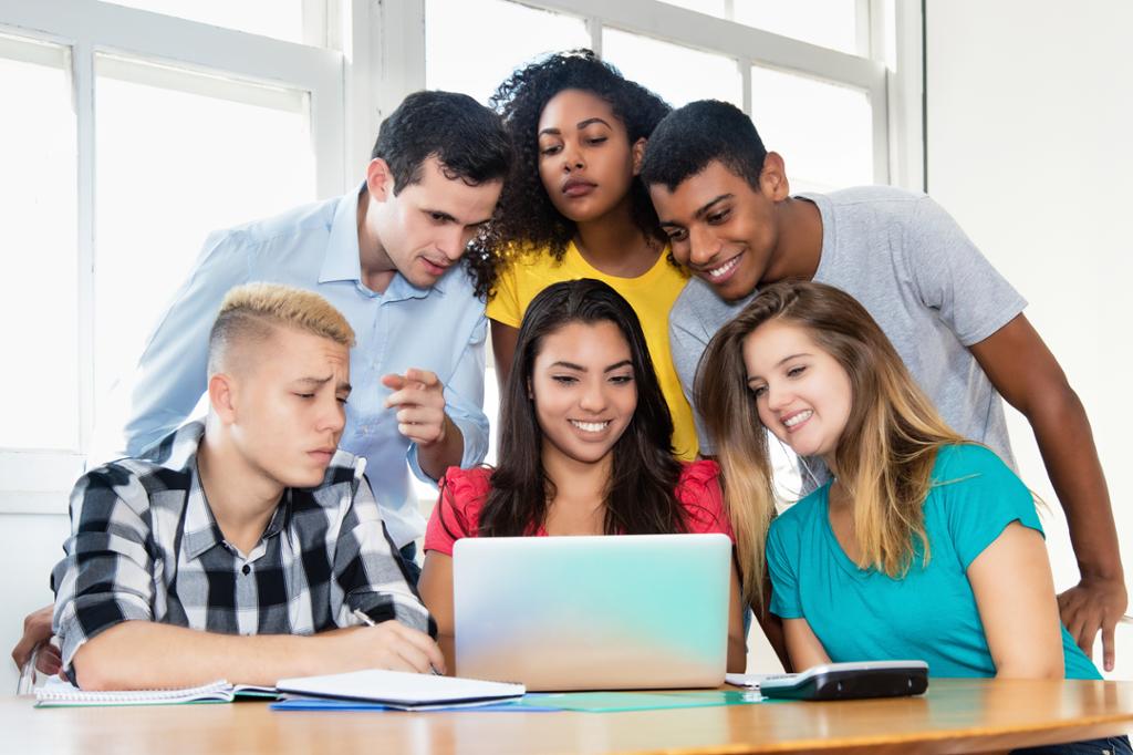Gruppe av studenter oppdaterer seg på dagens nyhetsbilde. 6 personer står sammen i en gruppe rundt en datamaskin.