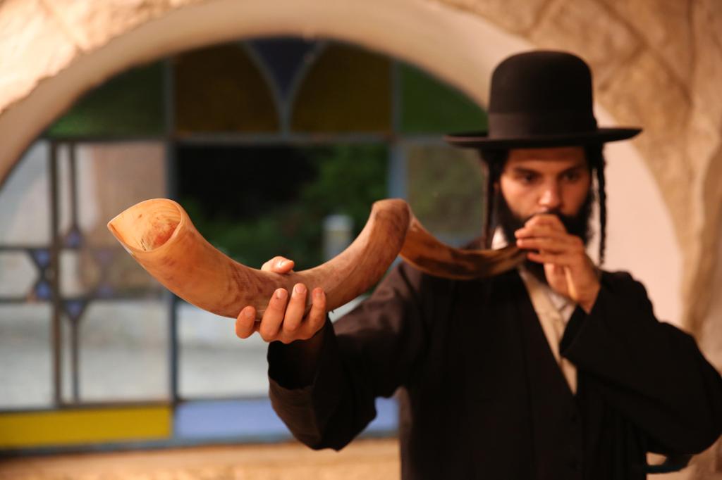Oultraortodoks jødisk mann blåser i bukkehorn (shofar). Mannen er iført svart dress og hatt, og har lange krøller ved ørene. Foto