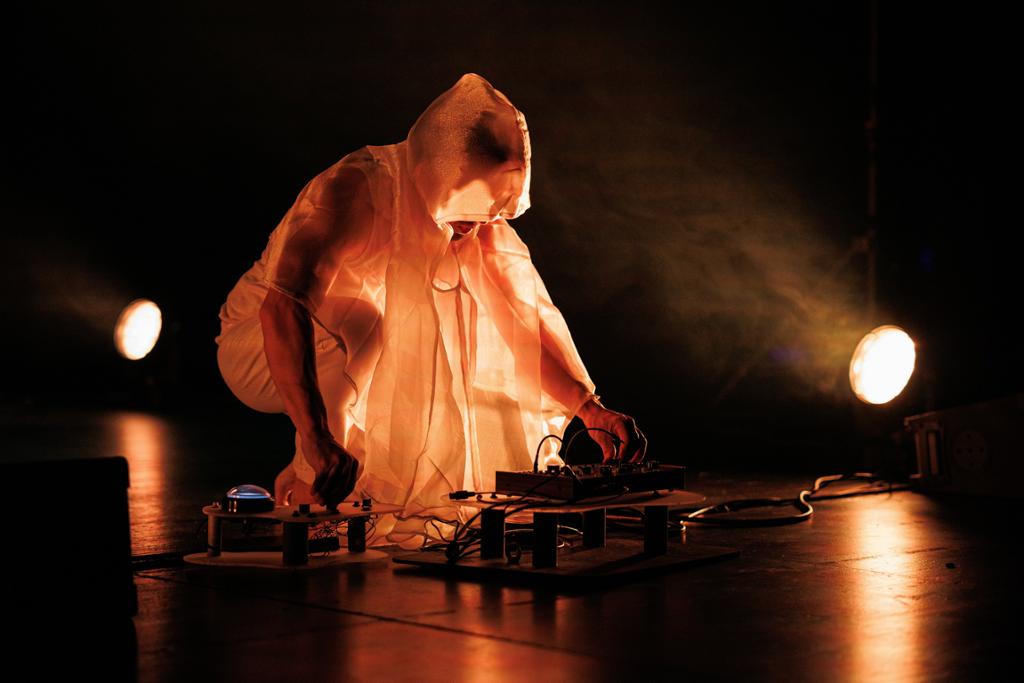 Person kledd i lyst slør sitter bøyd over en synthesizer. Scenen er mørk, men personen er opplyst av to lyskastere. Foto. 