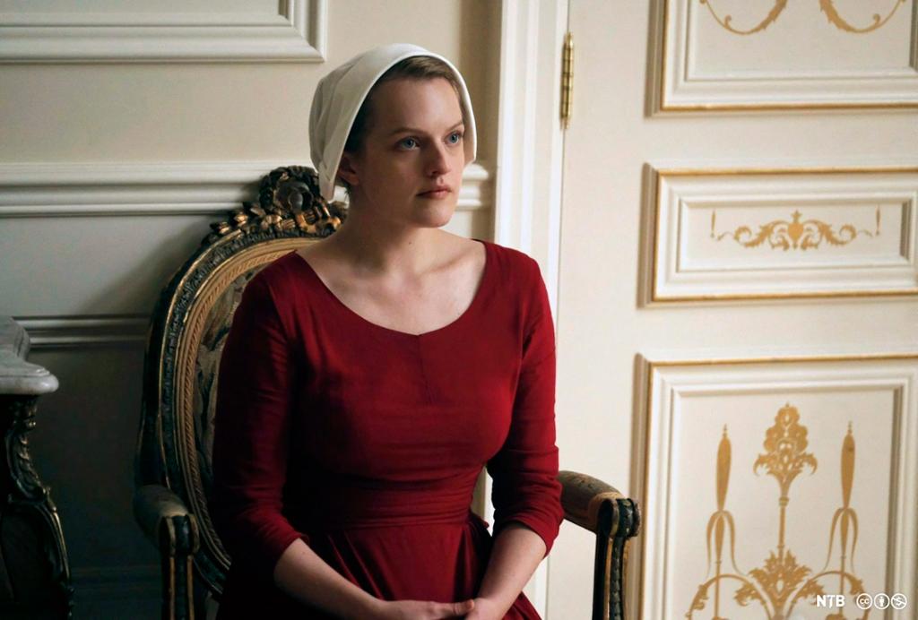 Tjenerinnen i TV-serien The handmaid's tale sitter i en stol med hendene i fanget og oppmerksomt blikk, iført rød kjole og hvit hette. Foto.