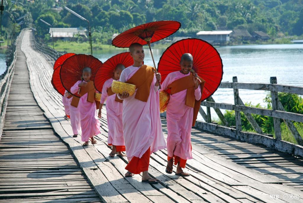 Smilende nonner med rosa kjortler, rød paraply og krukker i den ene handa går over ei bro. Foto.