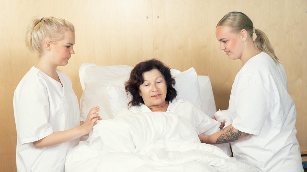 To helsefagarbeidere står på hver sin side av en sengeliggende pasient. Foto.