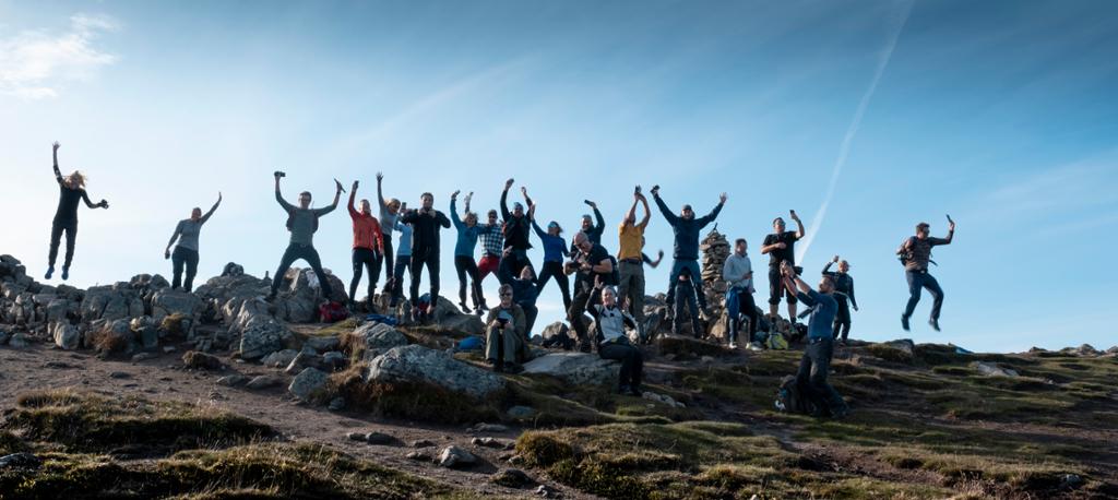Ei gruppe personar på ein fjelltopp hoppar og strekker armane i veret. Foto.