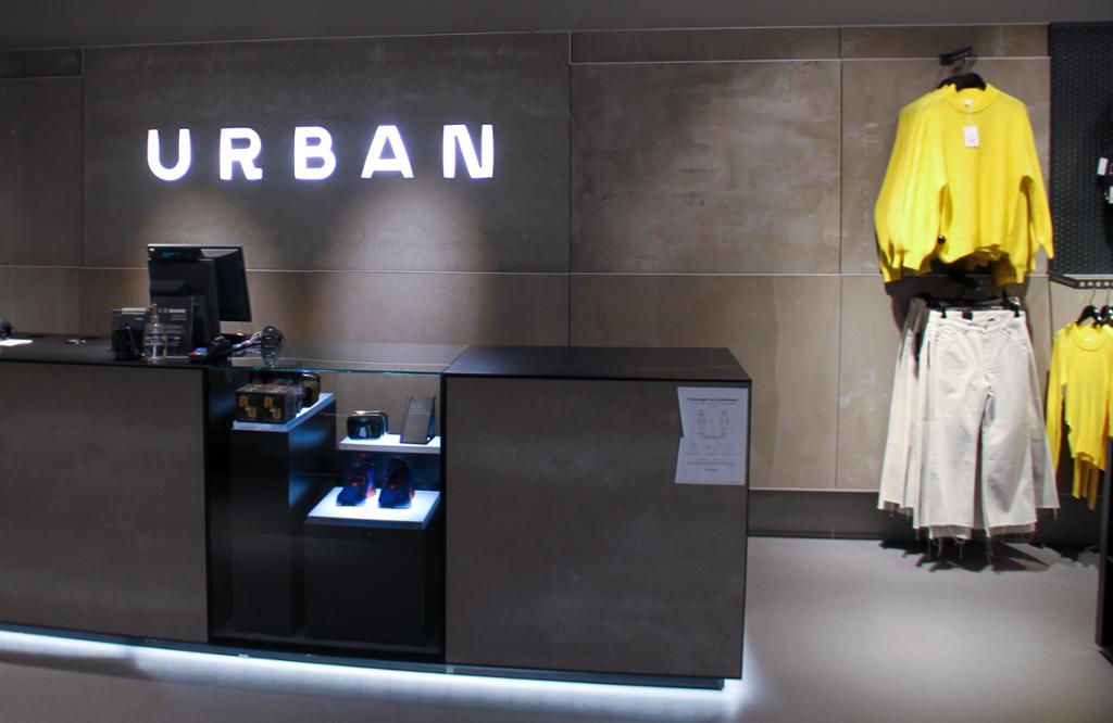 Kasseområdet til klesbutikken Urban på et kjøpesenter. Logoen til kjeden Urban er laget med lysende bokstaver og henger på veggen bak kassa. Foto.