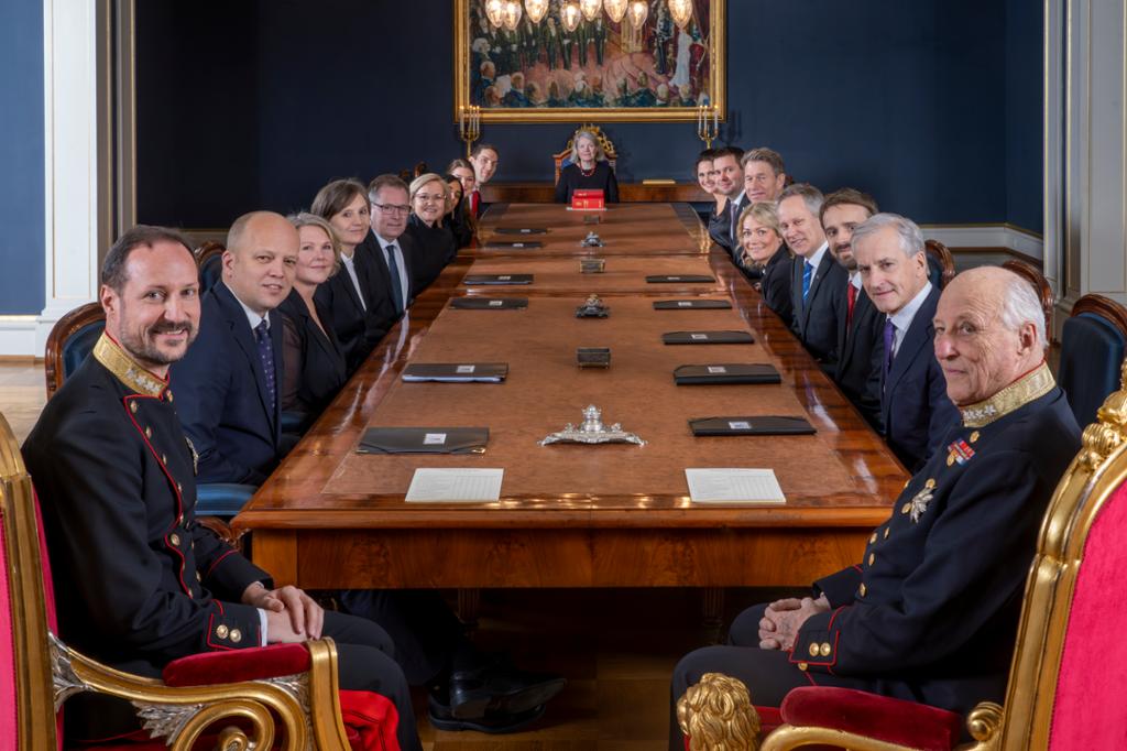 Støre-regjeringa sit ved eit bord saman med kronprins Haakon og kong Harald. Foto.