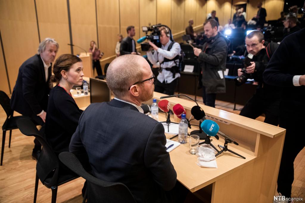 To menn og en dame ved et bord med mikrofoner foran mange journalister med kamera. Foto.