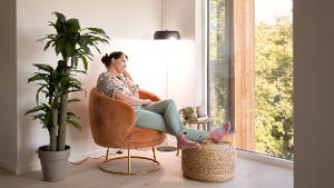Kvinne sitter i en  stol og ser ut et vindu. Lampe lyser i bakgrunnen. Foto.