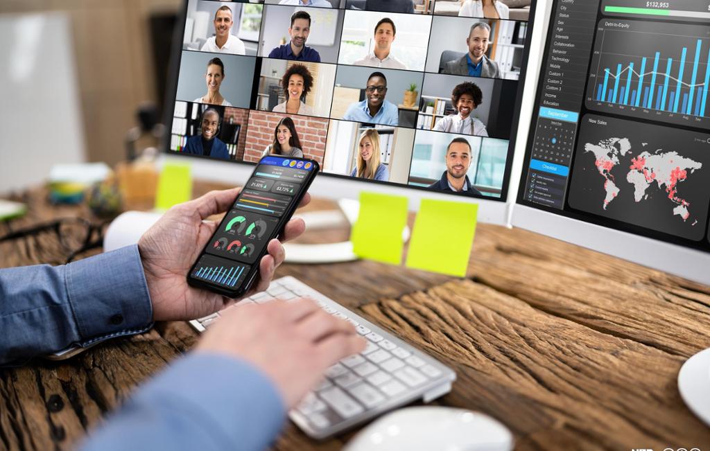 Person holder en mobil med grafikk og sitter ved en arbeidsplass med to dataskjermer. På den ene skjermen vises bilder av ulike personer, og på den andre skjermen vises et stolpediagram og et verdenskart. Foto.