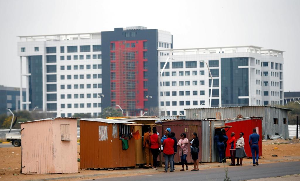 Modern building behind sheds. People in a line outside the sheds. 