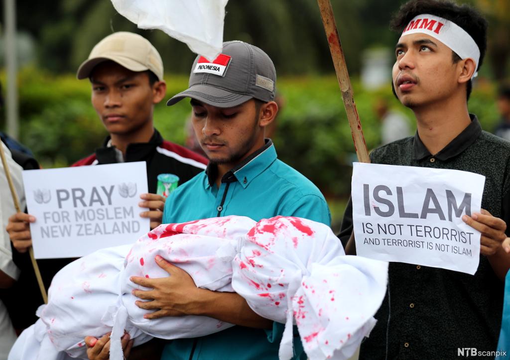 Tre menn med plakatar og ein blodflekt bylt i armane. På den eine plakaten står det på engelsk: "Islam er ikkje terrorisme, og terrorisme er ikkje islam". Foto.