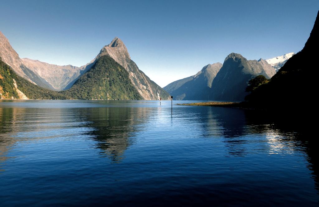 Photo: We see a fjord with steep mountains. The sky is blue. 