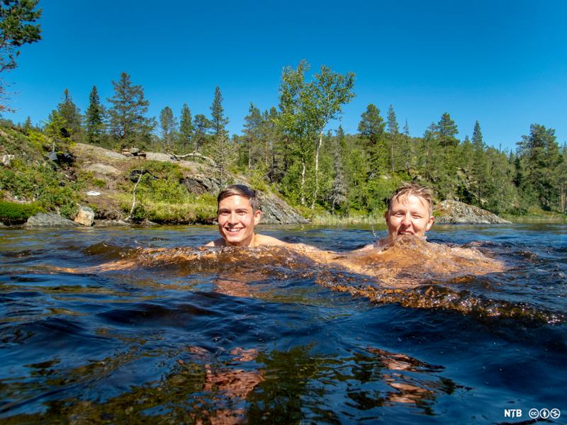 To gutter på atten til tjue år bader i et lite skogsvann. Begge smiler bredt mot kamera. Himmelen er skyfri.