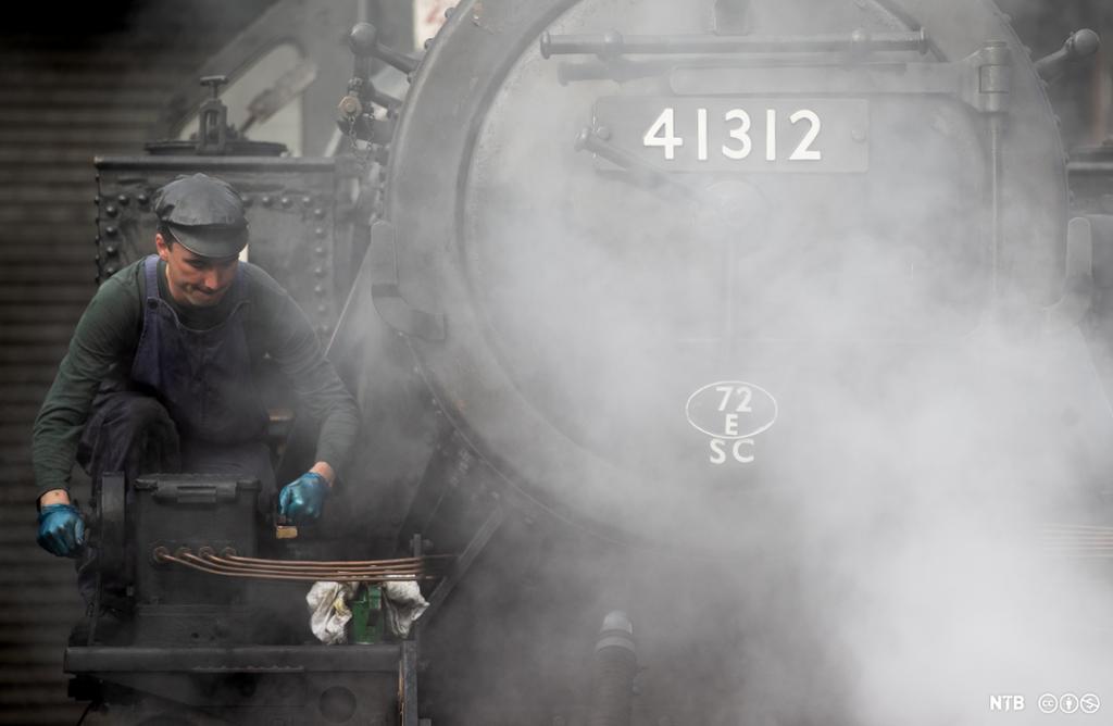 Mann med hatt, mørkt arbeidstøy og hanskar står ved sida av eit eldre damplokomotiv. Foto.
