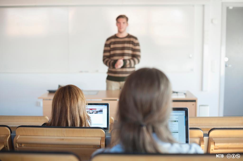 Bakhodet til to studenter med hver sin datamaskin foran seg. I bakgrunnen står en lærer. Foto. 
