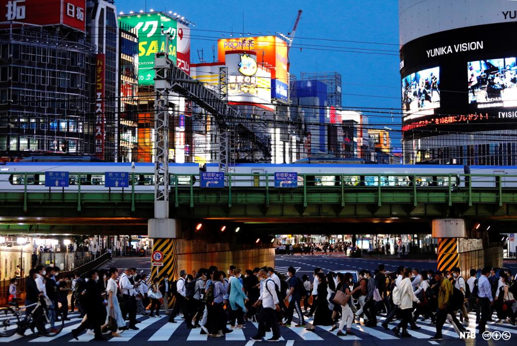 En mengde mennesker krysser en vei i Tokyo. Vi ser tog som passerer, og mange lyssatte bygninger. Foto. 
