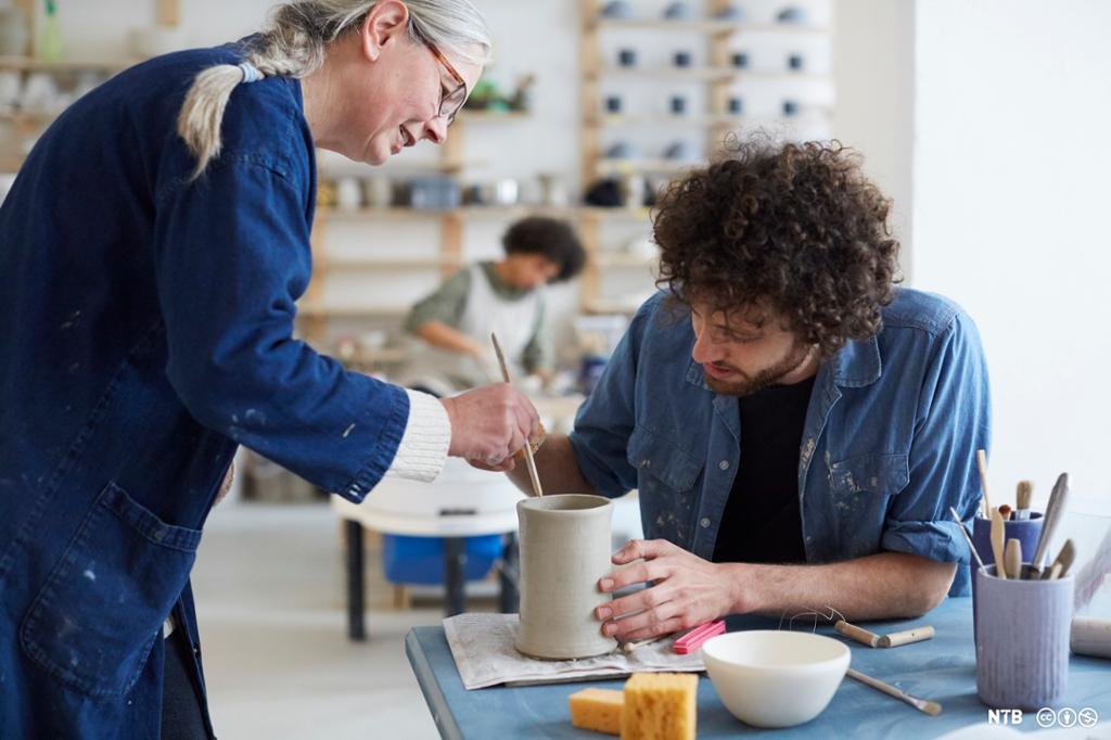 Kvinne og mann på ein verkstad; dei jobbar med ein keramikkvase som ikkje er brend enda. Foto.