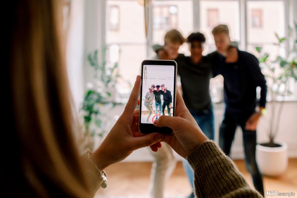 Jente med mobiletelefon tar bilde av tre gutter som poserer sammen. Foto. 
