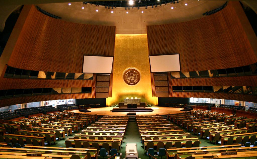 Stor sal med scene, mange bord med sete vender mot scenen. Rommet er rundt med mørke veggar. På scenen er det eit bord med tre sete og eit forgylt område med FNs logo på bakveggen. Foto.