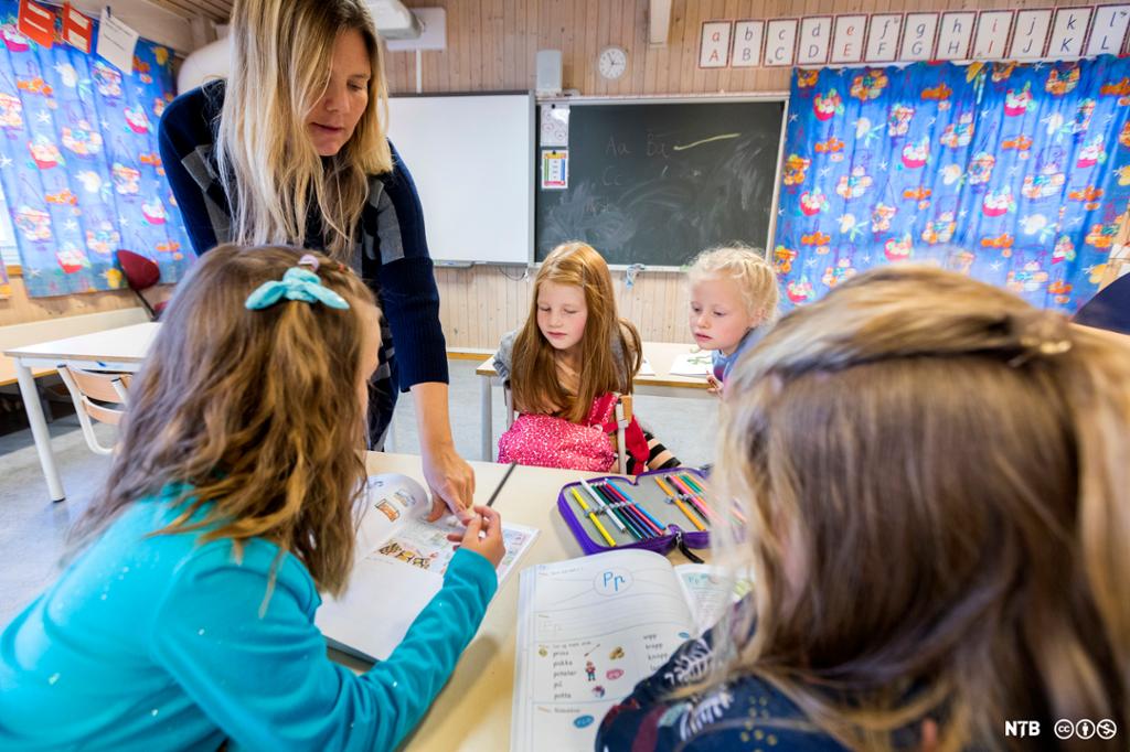 Kvinneleg lærar hjelper fire barneskuleelevar i eit klasserom. Ho peiker på noko i boka til den eine jenta. Foto.