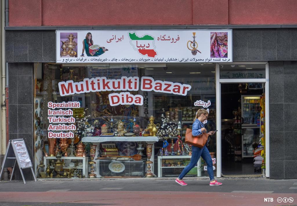Fotografi som viser butikkvinduet til en innvandrerbutikk i Köln i Tyskland. Over vinduet er det et stort reklameskilt som blant annet viser et kart over Iran i landets farger, og en kvinne som holder et fat med mat. På selve butikkvinduet står det: Multikulti-basar Diba, iranske, tyrkiske, arabiske og tyske spesialiteter. 