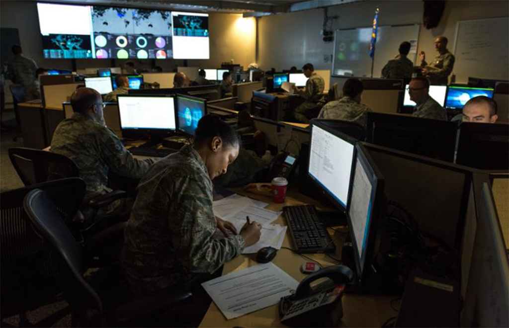 Soldater i militæruniform sitter foran dataskjermer i et kontorlandskap. De har utskrifter på ark liggende på pultene foran seg. Foto.