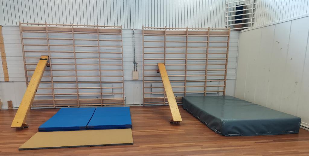 To benker står skrått opp mot ribbeveggen i en gymsal. Mellom benkene ligger det matter og en tjukkas. Foto.