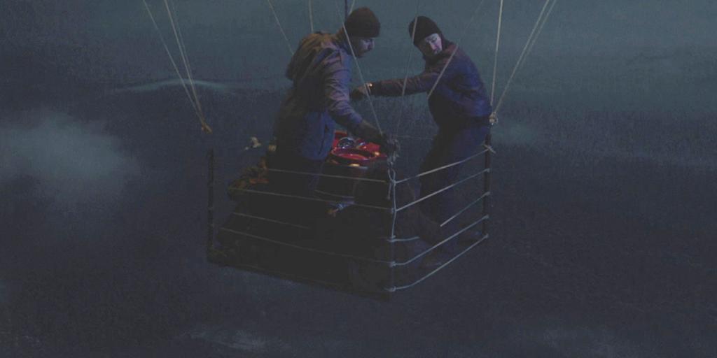 Utsnitt fra filmen "Flukten fra Øst-Tyskland": En varmluftsballong i lufta. I kurven er det to menn som står og ser nedover mot bakken, mens to andre sitter. Det er natt. Foto. 