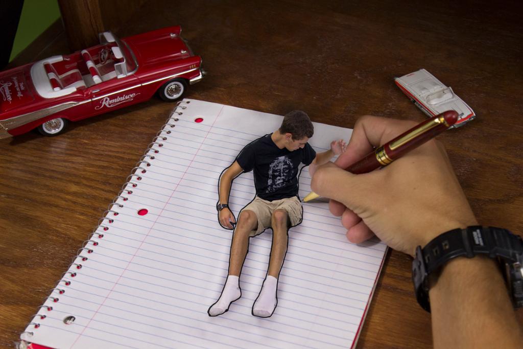 Picture of a hand drawing in a notebook, drawing an outline around a boy who sits up on the page of the notebook. There are toy cars on the desk. 