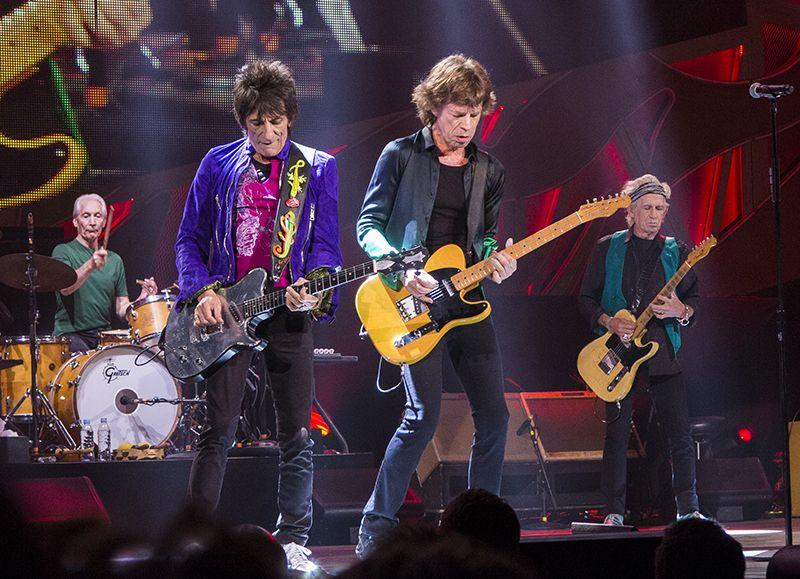Photo: The Rolling Stones in concert. We see three older men holding guitars, and one older man playing drums. 
