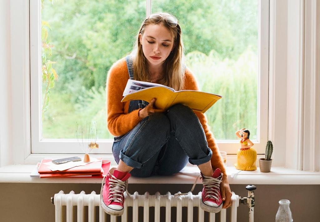 Jente studerer tekst i en bok. Hun sitter i en vinduskarm, har solbriller på panna, løse skolisser og beina på en oljefylt ovn. Foto.