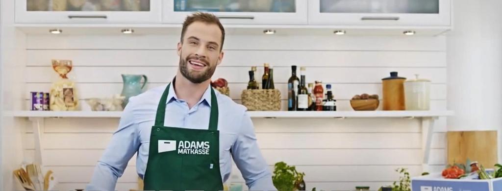 Adam Bjerck fra Adams Matkasse står på et lyst kjøkken og smiler mot kamera. Han har på seg lyseblå skjorte og et grønt forkle med firmalogoen til Adams Matkasse på. Foto.