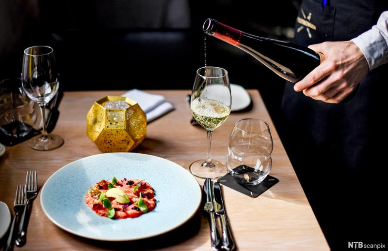 En tallerken med gourmetmat på et pent dekt bord. Hånden til en kelner sjenker hvitvin i et glass ved tallerkenen. Foto.