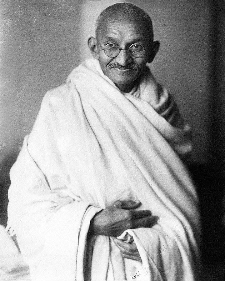 Black and white photo of Mahatma Gandhi. He's wrapped in a white cloth and wearing glasses. He's looking directly into the camera, smiling.   