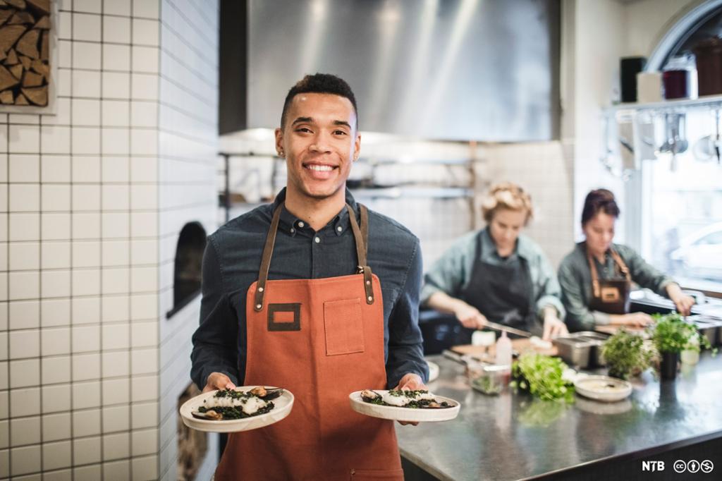 Smilende mannlig servitør med oransje forkle holder to tallerkener. I bakgrunnen ser vi to kokker på kjøkkenet. Foto.